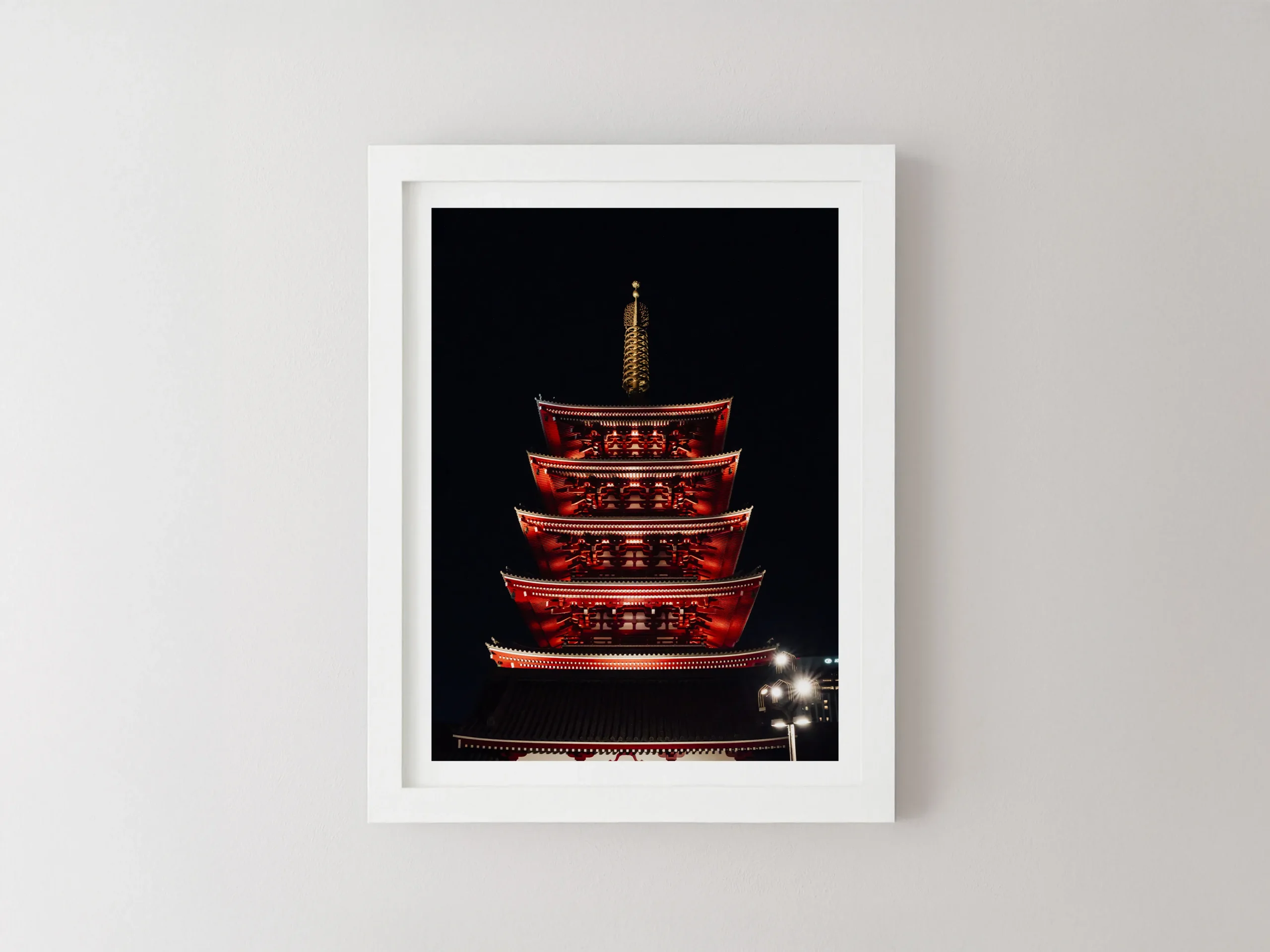 Tirage photographique d'art encadré de blanc, suspendu sur un mur texturé, représentant une pagode japonaise rouge vif à plusieurs niveaux, intensément éclairée contre un ciel nocturne noir de jais. La structure complexe est le point central, avec un finial doré au sommet.
