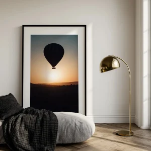 Grand cadre noir montgolfière posé au sol dans un intérieur minimaliste avec lampe dorée.