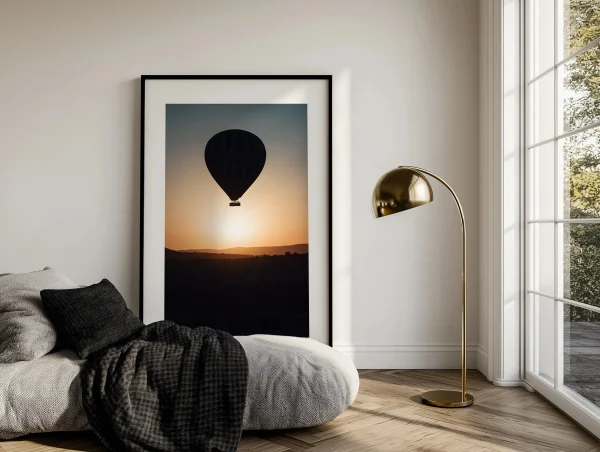 Grand cadre noir montgolfière posé au sol dans un intérieur minimaliste avec lampe dorée.