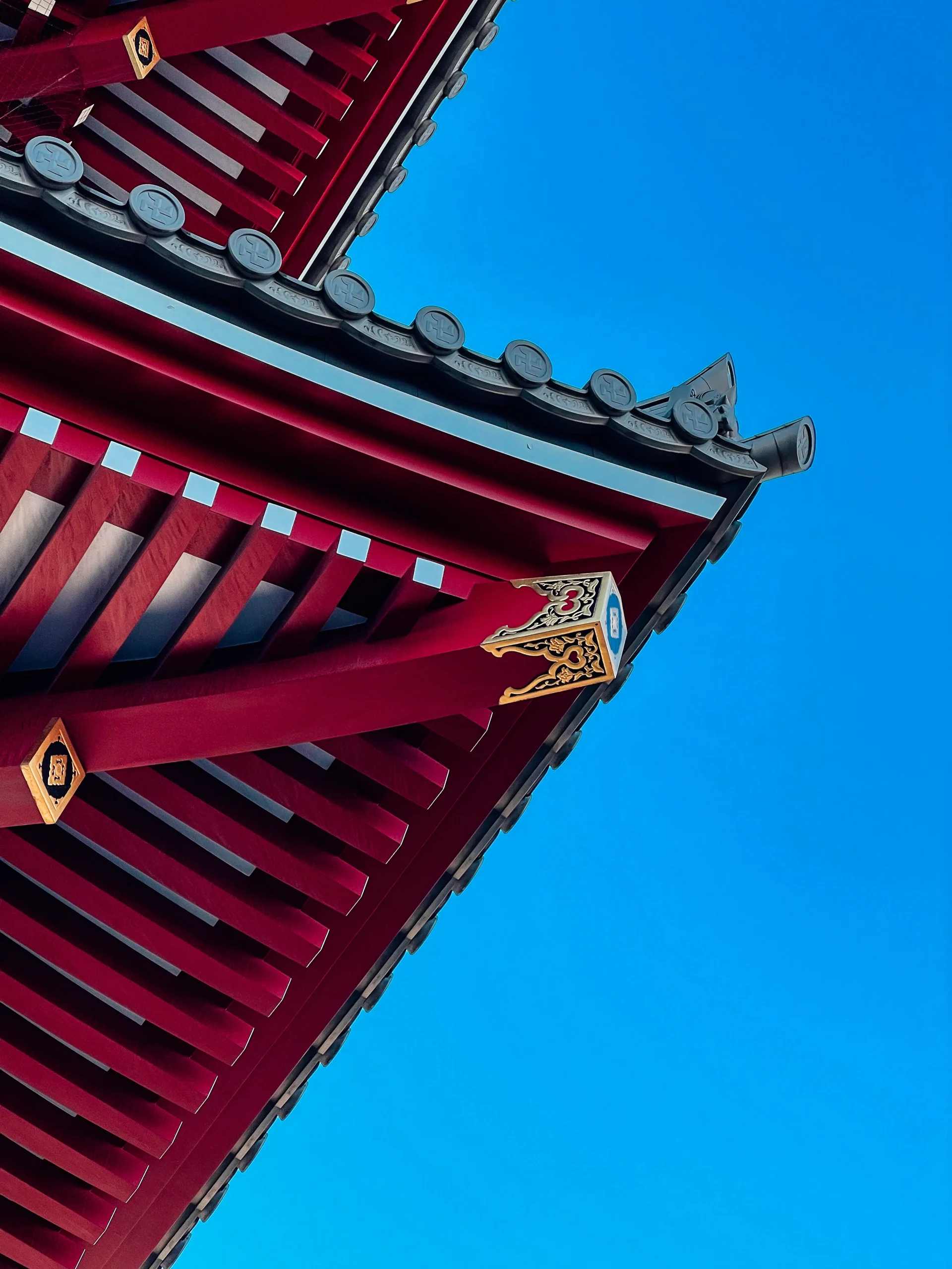 Photographie d'art minimaliste mettant en valeur l'architecture rouge et les boiseries d'un toit de temple traditionnel à Tokyo, sur fond de ciel bleu.